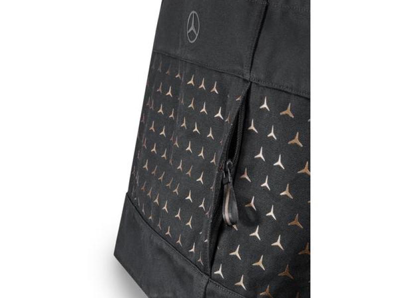 Nahaufnahme einer schwarzen Mercedes-Benz Einkaufstasche mit dreieckigem Muster in Bronze und Weiß, inklusive Mercedes-Logo auf der oberen Seite. Das Detailbild zeigt eine Reißverschlusstasche, die Teil des Designs ist.